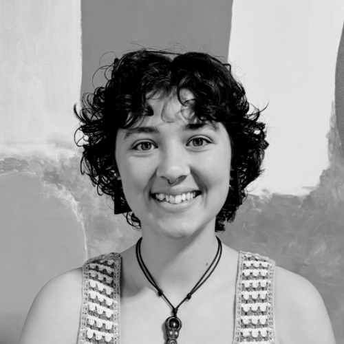 Smiling young woman with short curly hair, wearing a crocheted sleeveless top and pendant necklace, stands before an abstract mural.
