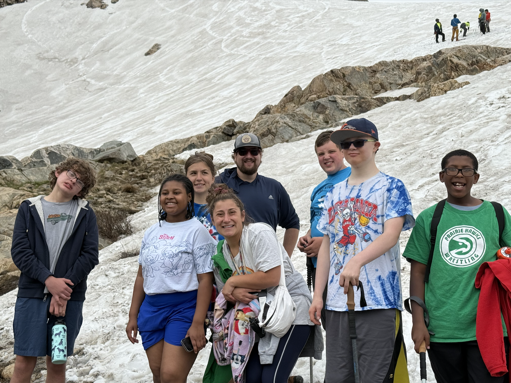 Group of smiling kids and teens posing on a snowy mountain slope with rocky outcrops, wearing hats and backpacks on a field trip. A few hikers are visible in the background.